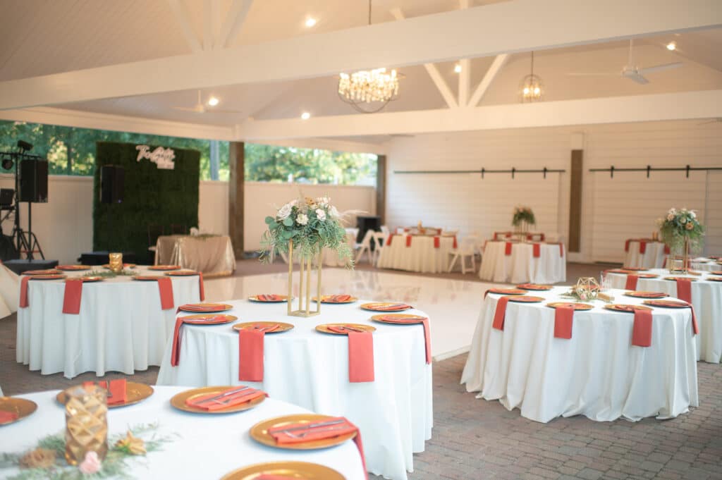 outdoor fall weddings at this venue are set with White tables and topped with a gold decorative plate charger and a coral napkin is draped on top of the gold charger held down with a silver for and knife. The tables are topped with gold stands that hold greenery, white roses and burnt orange roses. A white dance floor is centered underneath a large, circular chandelier that is dripping with crystals. A sweetheart table is the focal point of the reception area. A greenery wall with a neon sign is the backdrop to a table with a rose gold gold sequin tablecloth over a rectangle table. A "Mr. and Mrs." sign sits on top of the table. The wedding reception is all set underneath a large white pavilion that has a wall with shut, white sliding barn doors. The tables are strategically set for the perfect wedding experience for a Texas wedding. 