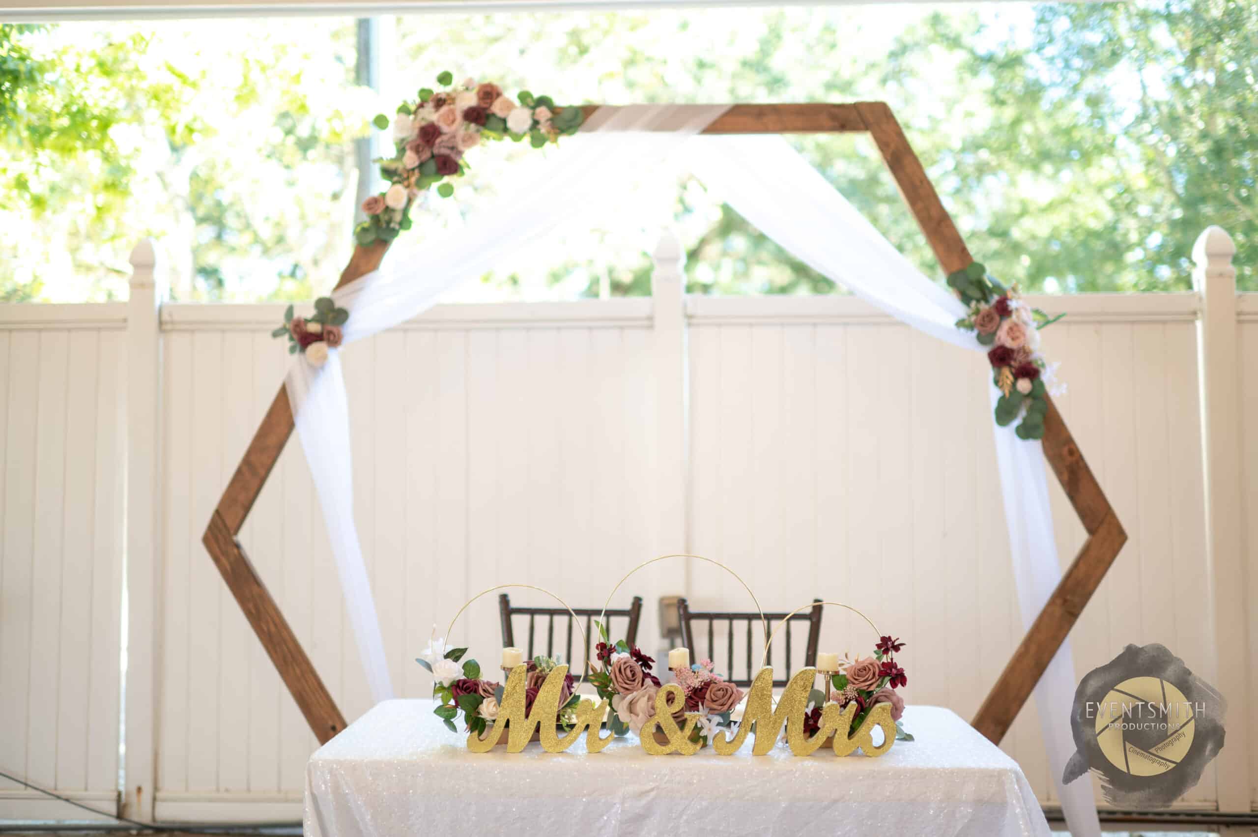 A wooden hexagon is decorated with white drapery hanging across the tops and down the sides. It sits behind a white tables where the bride and groom will sit known as a sweetheart table. There are wedding flowers decorating the wooden hexagon and then white table in front. A gold Mr & Mrs sign sits on top of the table. .