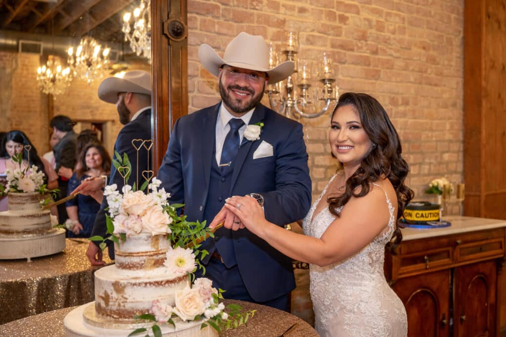 Bride in lace wedding gown has her hand with her wedding ring placed over the groom's hand while he holds a gold knife to cut wedding cake. Groom is in a navy suit with a white rose pinned to jacket and a beige cowboy hat. The wedding cake is a naked cake with gold designs sprayed onto the cake. Blush roses and greenery cascade down the 3 tier cake. 4 gold hearts at varying heights top the cake. The cake sits on a white cake stand with a rose gold sequin tablecloth underneath it. Bride and groom are smiling and looking at the camera. 