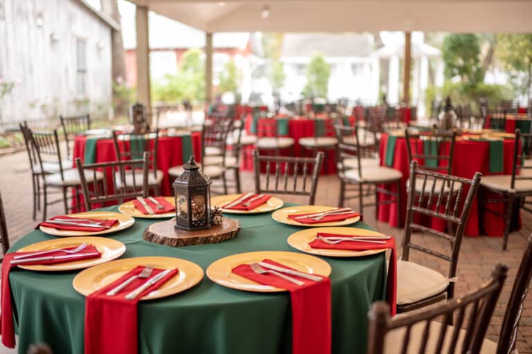 Outdoor Christmas Wedding/Party setup. Emerald table linen with red napkins sitting on top of a gold plate charger. Black lantern has pinecones in it and sits on a wooden stump.