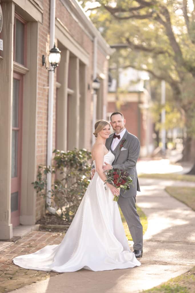 Bride and Groom smiling under the historic oaks of League City in front of brick building that is an industrial chic historic wedding venue Butler's Courtyard