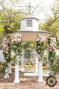 A one of a kind, white, wedding gazebo surrounded by meticulously curated landscaping sits beside a tranquil waterfall and pond. Adorned with custom, Lush Flowers floral arrangements. The Butler's Courtyard gazebo exudes beauty and elegance, creating a picturesque and enchanting setting, making it the most popular wedding ceremony location near Houston, Texas.