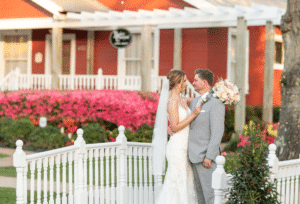 A smiling bride and groom stand on a charming bridge, surrounded by vibrant spring landscaping in full bloom. Behind them, a row of bright pink azaleas adds a splash of color, perfectly capturing the essence of a beautiful spring wedding scene at Butler's Courtyard, a stress free wedding venue near Houston, Texas.