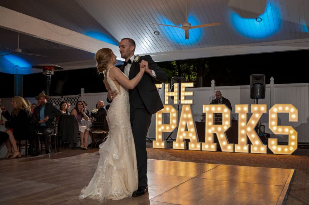 Bride and groom dancing in the outdoor pavilion with bright marquee letters spelling their last name, showcasing unique lighting and customized dance floor backdrop at Butler's Courtyard, a full-service Houston wedding venue.
