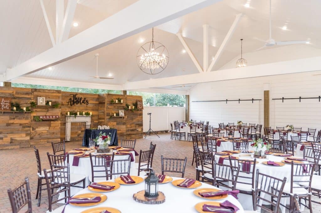 Wedding reception tables set under the wedding pavilion at Butler's Courtyard, a premiere wedding venue Houston Texas, with a rustic wood wall backdrop, featuring maroon and gold décor because the couple are Texas A&M Aggies.