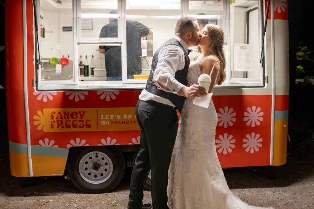 Bride and groom share a kiss in front of a sno cone truck, highlighting unique wedding trends and memorable moments that can be created at Butler's Courtyard.
