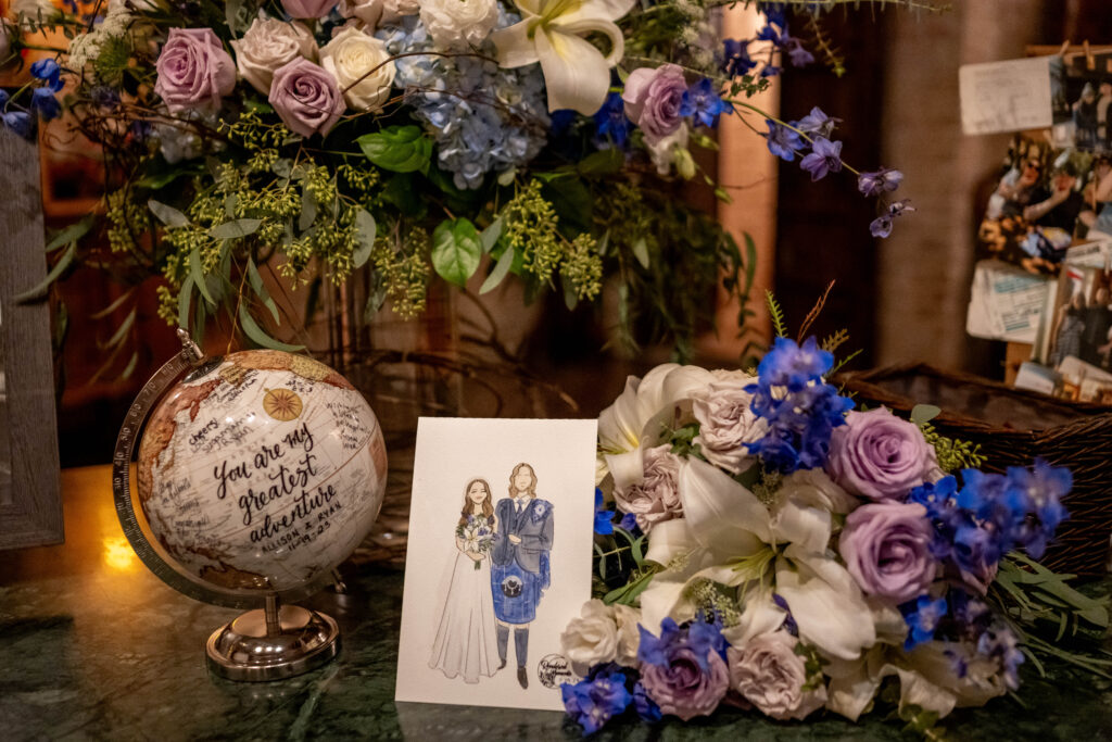 Wedding entry table at Butler's Courtyard with blue and purple flowers beside a globe and watercolor painting of the bride and groom, showcasing 2025 wedding trends at a historic venue with unique wedding ideas and elegant wedding flowers.
