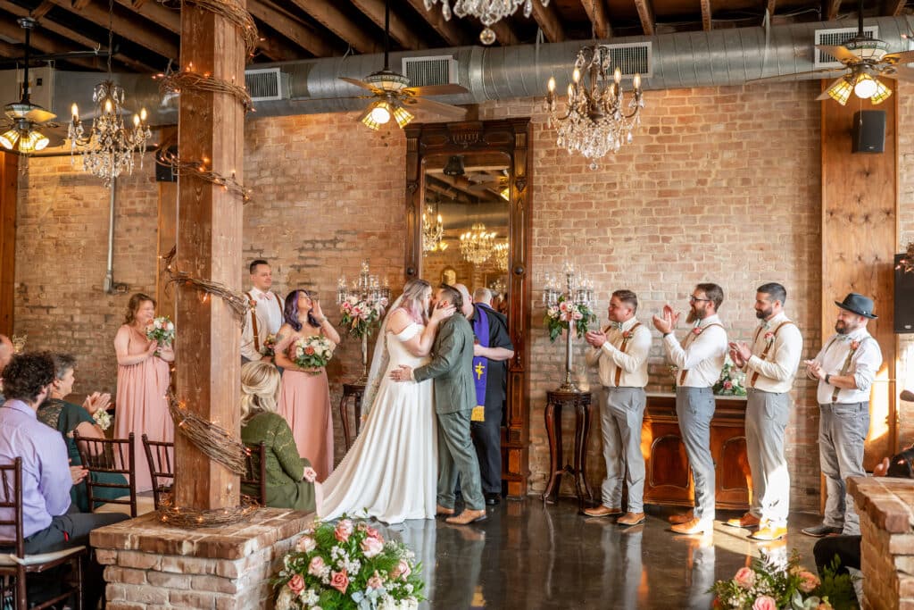 In the climate controlled ceremony location known as the Grand Hall at Butler’s Courtyard near Houston Texas, a bride and groom share a romantic kiss at the end of their ceremony. The setting features historic brick walls elegantly decorated with chandeliers and candelabras, accompanied by beautiful blush florals, enhancing the romantic ambiance.