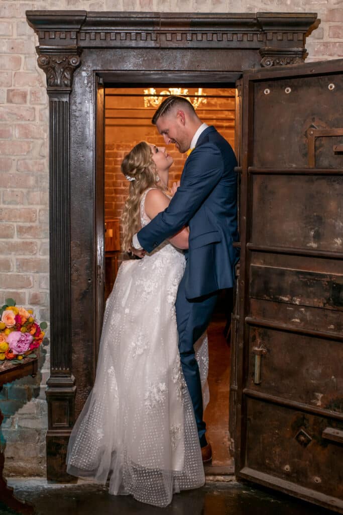 A bride in a stunning white wedding dress and a groom in a sharp navy suit share a romantic gaze while standing in the threshold of a historic bank vault in The City of League City. The timeless elegance of their attire contrasts beautifully with the vintage charm of the vault’s industrial chic design, capturing the unique allure of Butler’s Courtyard in League City as an unforgettable wedding destination blending romance and history.