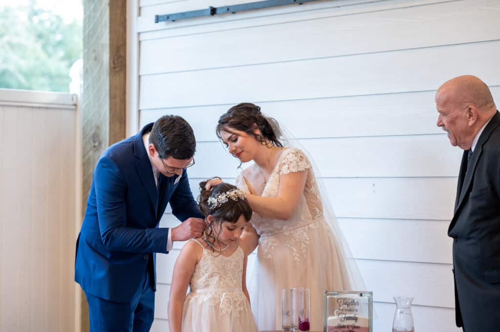 Bride and groom present their daughter with a keepsake necklace during their wedding ceremony at Butler's Courtyard, symbolizing their love and commitment to their united family.