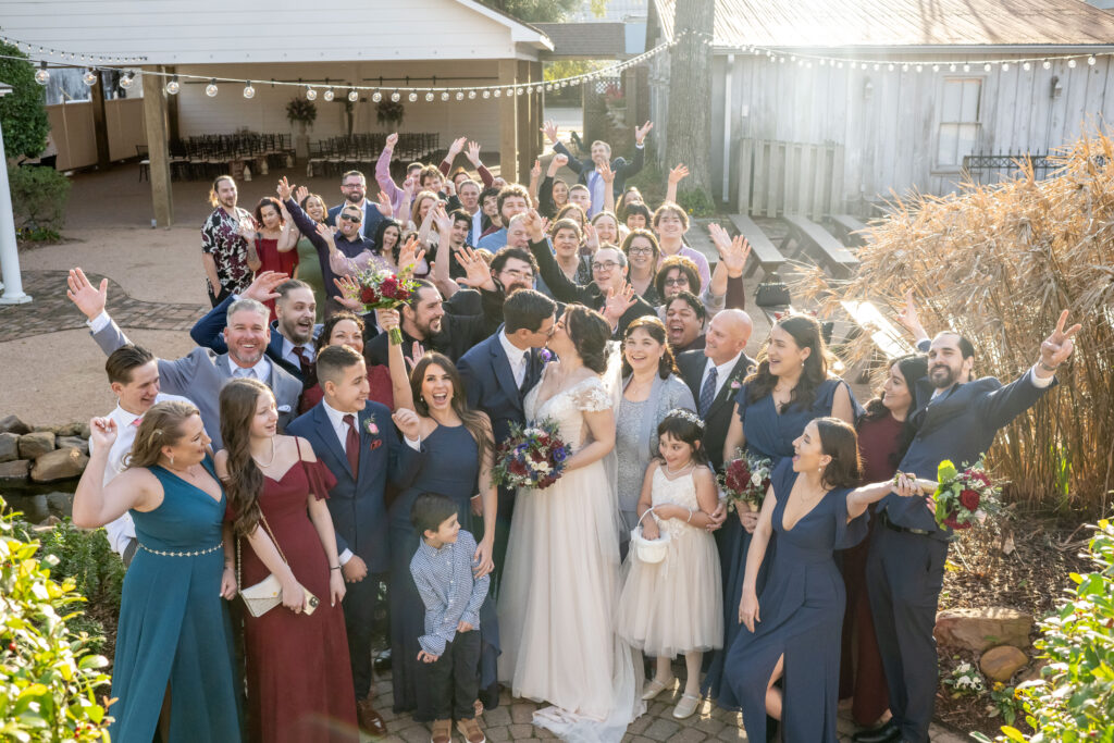 Bride and Groom kiss while their guests cheer and celebrate during the group shot taken at Butler's Courtyard featuring every guest in attendance. This is a once in a lifetime photo!