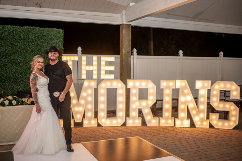 Happy Bride and Groom stand smiling on the dance floor in front of lighted marquee letters