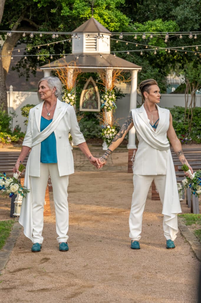 Two brides at their LGBTQ wedding in Houston, elegantly posing in front of a beautifully adorned wedding gazebo with custom teal and white florals, showcasing the charm of a garden wedding at Butler's Courtyard.