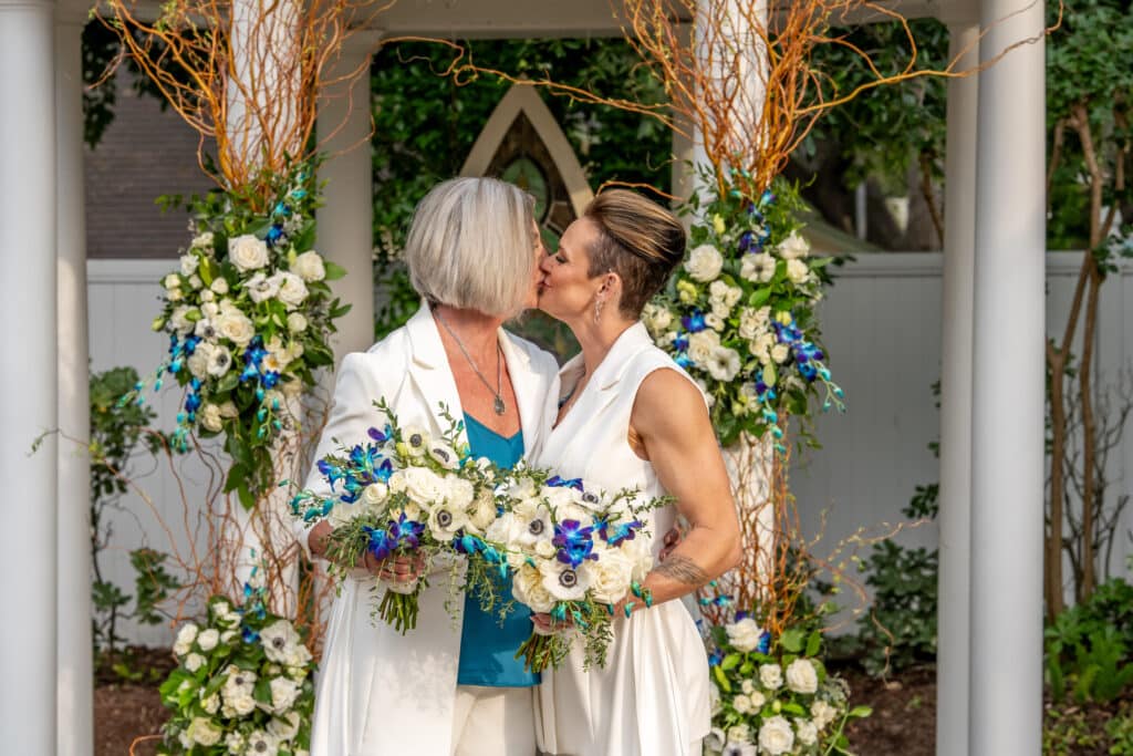 Two brides sharing a romantic kiss at their LGBTQ wedding in Houston, standing in front of a stunning wedding gazebo decorated with custom teal and white florals, highlighting the beauty of a garden wedding at Butler's Courtyard.