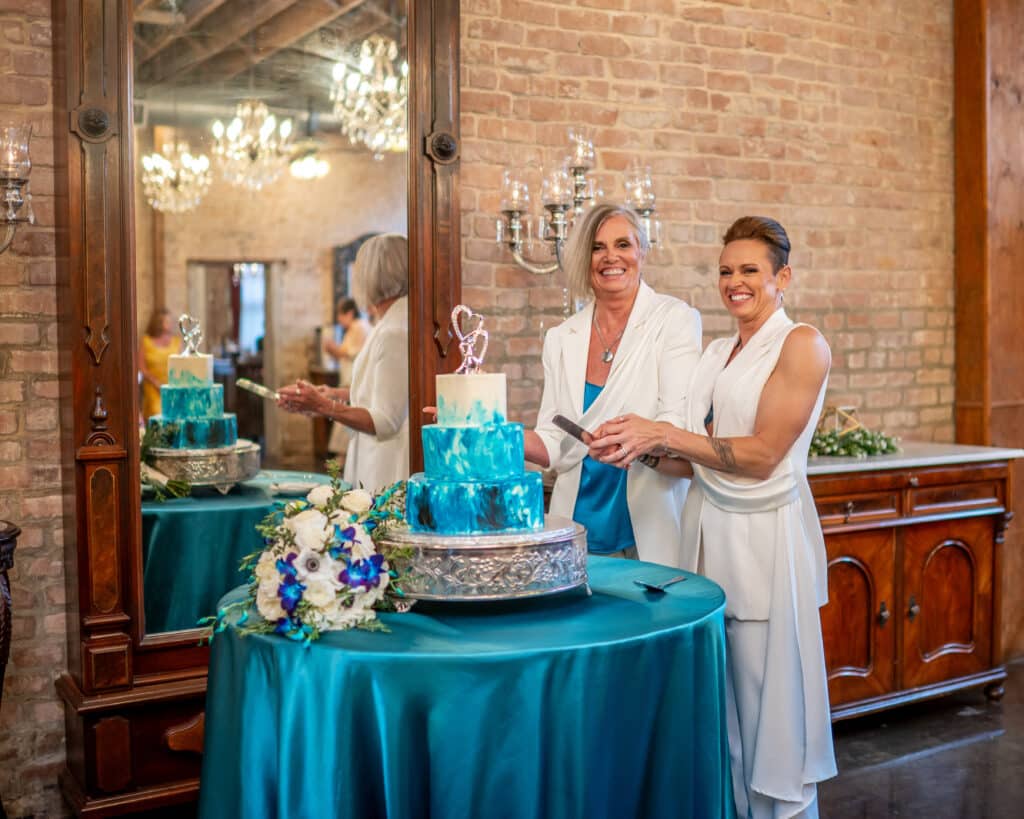 Two brides at their LGBTQ wedding in Houston, smiling brightly as they perform the ceremonial cake cutting. The cake showcases a striking teal ombre 'paint' effect, beautifully displayed on a table draped with teal linen, creating an elegant and joyful moment at Butler's Courtyard.