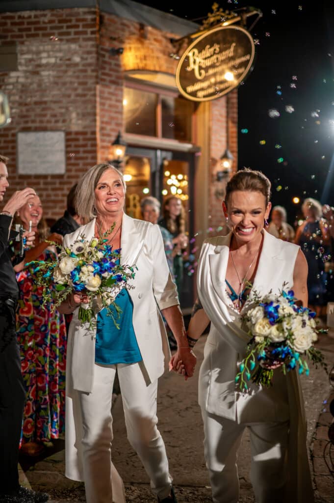 Two brides celebrating their LGBTQ wedding at Butler's Courtyard, wearing custom white suits with elegant teal accents, holding matching teal and white bouquets, and smiling joyfully as they walk through a cascade of bubbles during their grand exit.