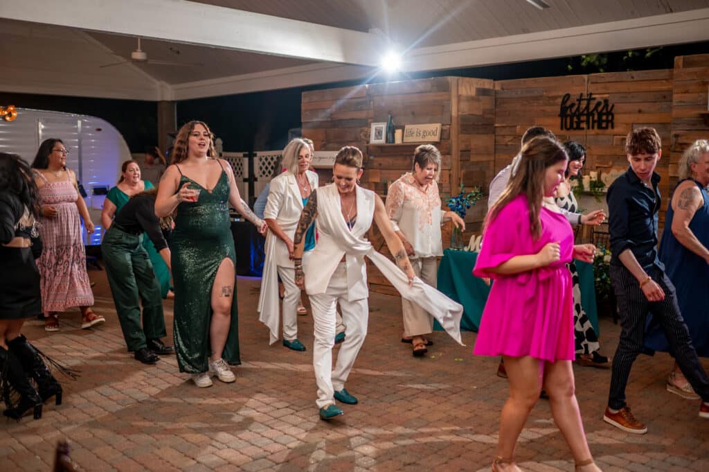 Two brides celebrating their LGBTQ wedding in Houston, dancing joyfully on a packed dance floor surrounded by smiling guests, capturing the vibrant and festive atmosphere of a wedding at Butler's Courtyard.