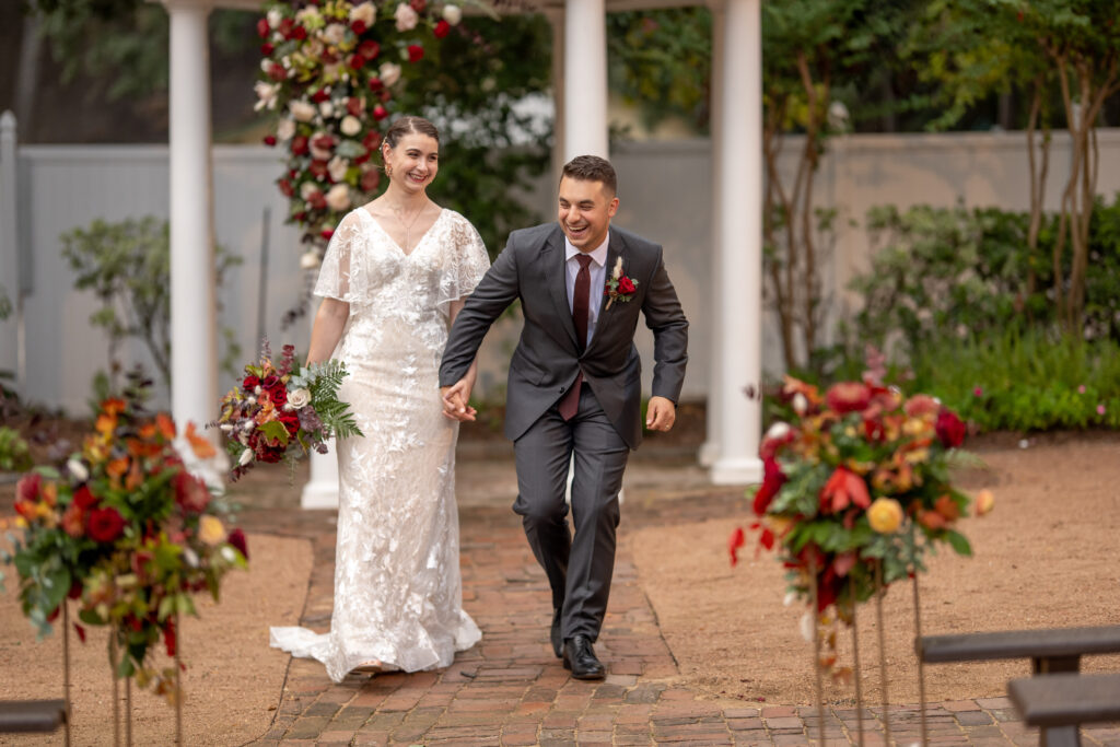 Bride and groom smiling and laughing during their outdoor fall wedding in Houston at Butler's Courtyard, surrounded by vibrant fall-colored florals showcasing a warm and festive atmosphere perfect for autumn celebrations.